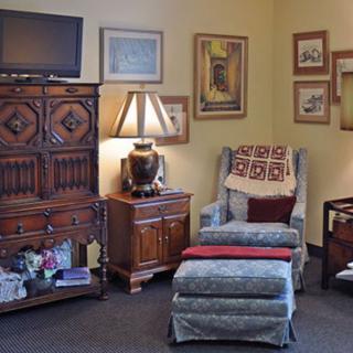 A cozy living room with an armchair and ottoman, decorated with a lamp, cabinet, and various wall art.