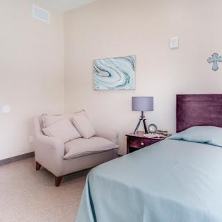 A cozy bedroom featuring a bed, an upholstered chair, and a nightstand with a lamp.