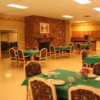An empty dining hall with neatly arranged tables and chairs, featuring a fireplace and adjacent kitchen area.
