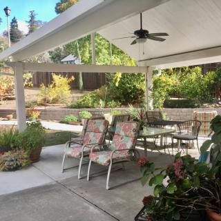 A cozy outdoor patio with chairs and a table under a covered area, surrounded by lush garden plants.