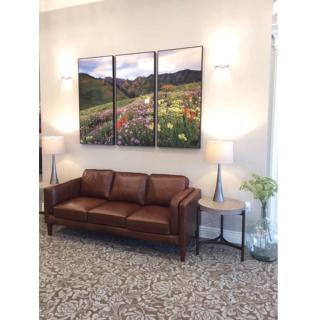 A cozy living room setup featuring a brown leather sofa, modern lamps, and a three-panel landscape painting.