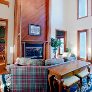 A cozy living room with a plaid sofa, wooden furniture, and a fireplace surrounded by large windows.