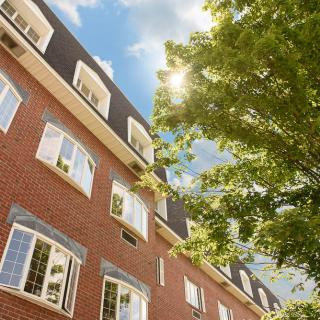 A brick building with numerous windows, surrounded by trees and basking in sunlight under a clear blue sky.