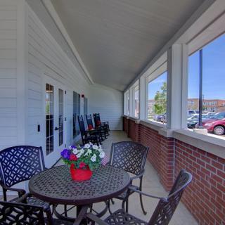 A cozy porch area with a table, chairs, and potted flowers, overlooking a parking lot.
