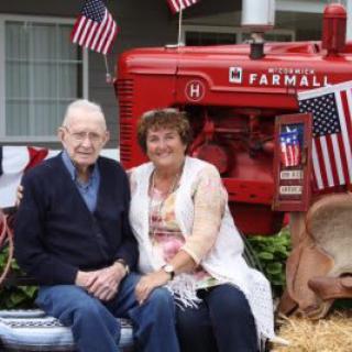 An elderly couple sitting in front of a red Farmall tractor decorated with American flags.