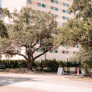 An outdoor scene featuring a large building surrounded by trees, with a fenced entrance and signage visible.