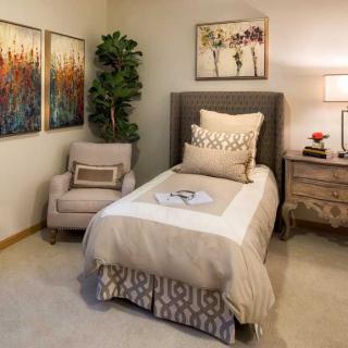 A cozy bedroom with a single bed, decorated in neutral tones, featuring an armchair, nightstand, and artwork on the walls.