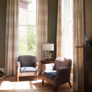 A cozy living room with two armchairs by a large window, adorned with curtains and a table lamp, basking in sunlight.