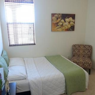 A cozy bedroom with a neatly made bed, artwork on the wall, and a patterned chair by the window.
