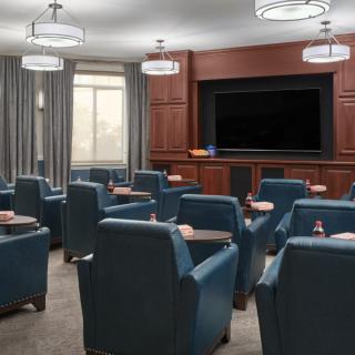 A modern meeting room with comfortable seating facing a large television, featuring wood paneling and elegant lighting.