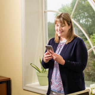 A woman standing by a window, engaged with her smartphone while a plant sits on the windowsill.