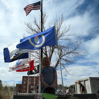 A person stands among various flags, including the American flag, against a backdrop of trees and a cloudy sky.