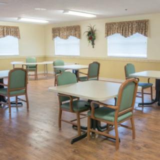 An empty dining area with multiple tables and chairs, featuring a wooden floor and windows with curtains.