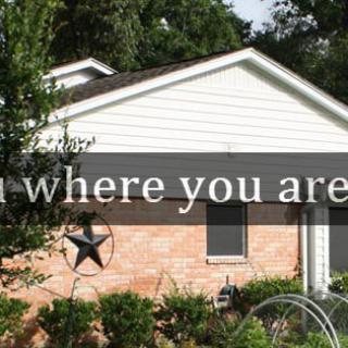 A brick house with a garden and decorative star, featuring the text 'Bloom where you are planted.'
