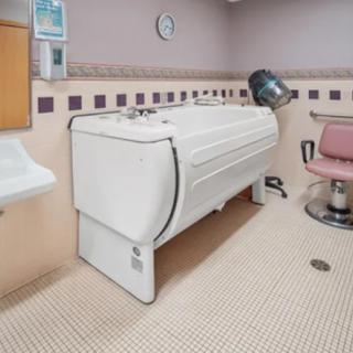 A healthcare facility room featuring a specialized bathtub and a salon chair equipped with a hair dryer.