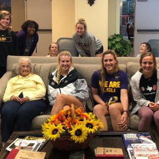 A group of people sitting and standing around a sofa in a lounge area, with flowers on the table.