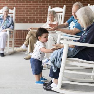 A young child interacts with elders on a porch filled with rocking chairs.