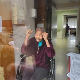 An elderly woman in a wheelchair joyfully gestures through a window, expressing happiness during a visit.