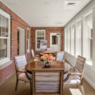 A cozy dining area with a brick wall, featuring a wooden table and chairs, adorned with flowers, and natural light streaming in through the windows.