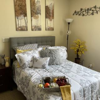 A cozy bedroom features a neatly made bed with patterned bedding, complemented by wall artwork and decorative elements like a floor lamp and plants. A tray with breakfast items is placed on the bed.