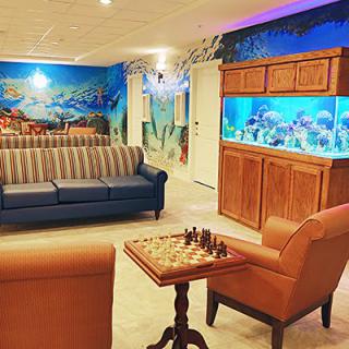 A cozy living space featuring a striped sofa, armchairs, a chessboard on a table, and a large aquarium set against an ocean-themed wall mural.