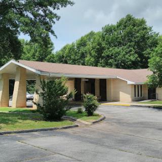 A single-story building surrounded by lush greenery, featuring a paved parking lot.