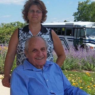 A smiling elderly man in a wheelchair sits in front of a woman with flowers and a bus in the background.