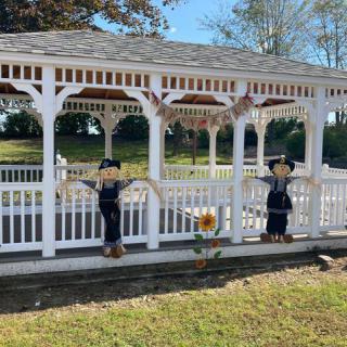 A white gazebo adorned with scarecrow decorations, set in a sunny outdoor park, capturing the essence of autumn festivities.