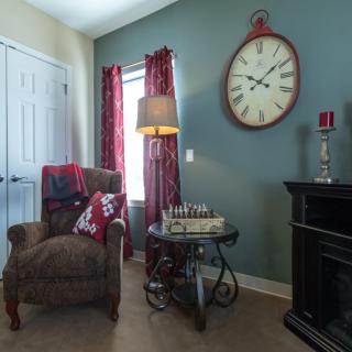 A cozy living room with an armchair and a side table topped with a chess set, illuminated by a lamp, featuring a large wall clock.