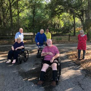 A group of people enjoying the outdoors, with some in wheelchairs, surrounded by trees and sunlight.