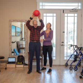 An elderly man participates in a physical therapy session, lifting a red exercise ball with the guidance of a therapist.