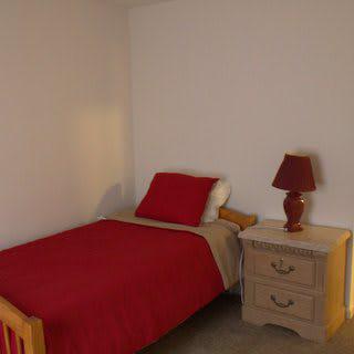 A minimalist bedroom with a single bed featuring red bedding and a nightstand with a lamp.