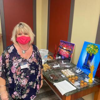 A woman wearing a floral shirt and a pink mask stands next to a table displaying cookies and artwork.