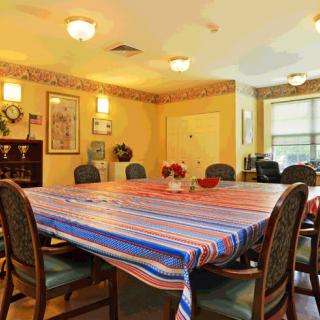 A well-lit dining room with a large table covered by a colorful striped tablecloth, surrounded by chairs.