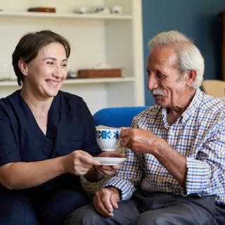 A caregiver enjoys a warm moment with an elderly man, sharing tea and laughter in a cozy living room.