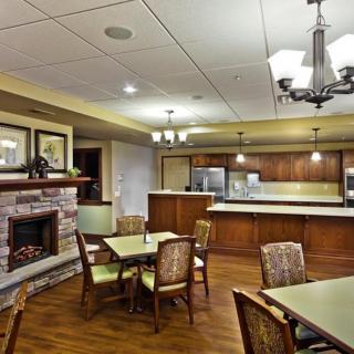 A cozy dining area with a fireplace and kitchen in the background, featuring tables and chairs under warm lighting.