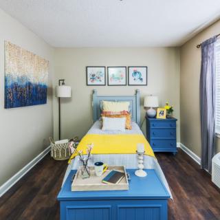 A cozy bedroom with a single blue bed and coordinated furniture, featuring framed artwork and a bright yellow accent throw.