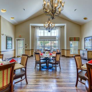 A well-lit dining room with wooden flooring and elegant chandeliers, featuring several tables and chairs arranged neatly.