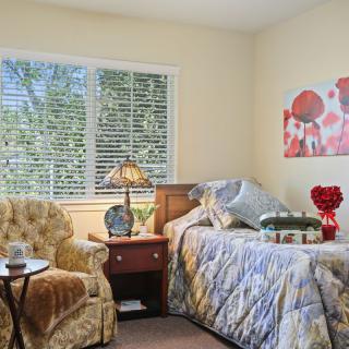 A cozy bedroom with a single bed, an armchair, and a nightstand under a large window.