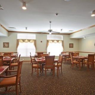 A well-lit dining room with multiple tables and chairs, featuring carpeted flooring and ceiling fans.