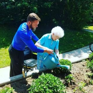 An elderly woman is being assisted by a man as they water the garden together.