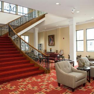 A spacious lobby featuring a grand staircase with red carpet, comfortable seating area, and large windows.