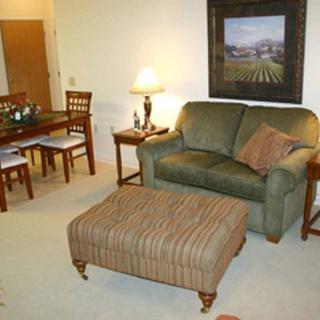 A cozy living room with a green sofa, striped ottoman, and a dining table in the background.
