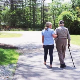 An elderly person walking with support from a companion in a serene outdoor setting.