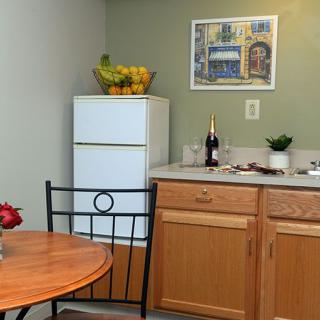 A cozy kitchen corner with a small table and chairs, featuring a fridge topped with a fruit basket, wooden cabinets, and a countertop adorned with a bottle and potted plant.