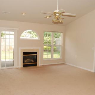 An empty living room with a fireplace, ceiling fan, and large windows.