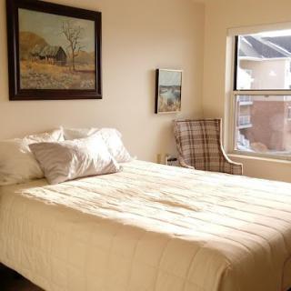 A cozy bedroom with a neatly made bed, an armchair by the window, and artwork on the walls.