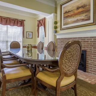 A cozy dining room with a glass table, upholstered chairs, and artwork above a brick fireplace.