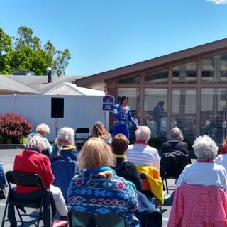 A community event taking place outdoors, with a speaker addressing a gathered audience of seniors.