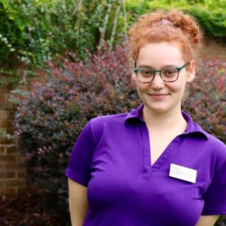 A person with red hair and glasses, wearing a purple shirt with a name tag, smiling in a garden.